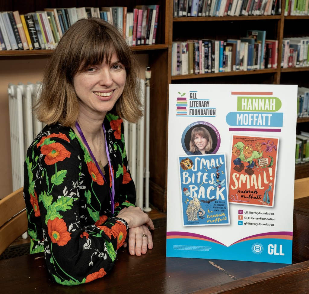 Me, in a black dress with red poppies at a desk next to a sign with a photo of me and the covers of Small and Small Bites Back